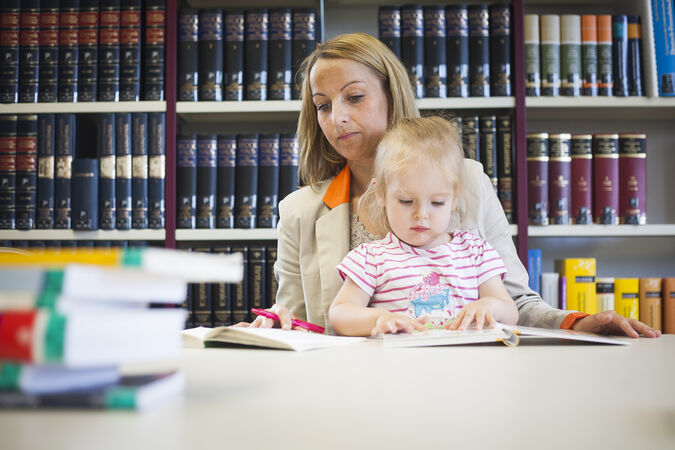 Eine Mutter sitzt mit ihrem Kind auf dem Schoß in der Bibliothek der Hochschule. Die Mutter hat vor sich einen Stapel Bücher und schreibt. Das Kind blättet in einem Bilderbuch.